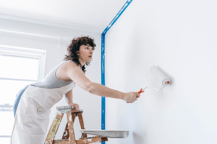 Femme sur un escabeau passant un rouleau de peinture blanche sur un mur, avec un ruban bleu de masquage
